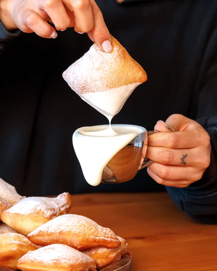 Một bàn tay nhúng chiếc beignet phủ đường bột vào tách kem trắng nhỏ, phía trước là khay beignet.