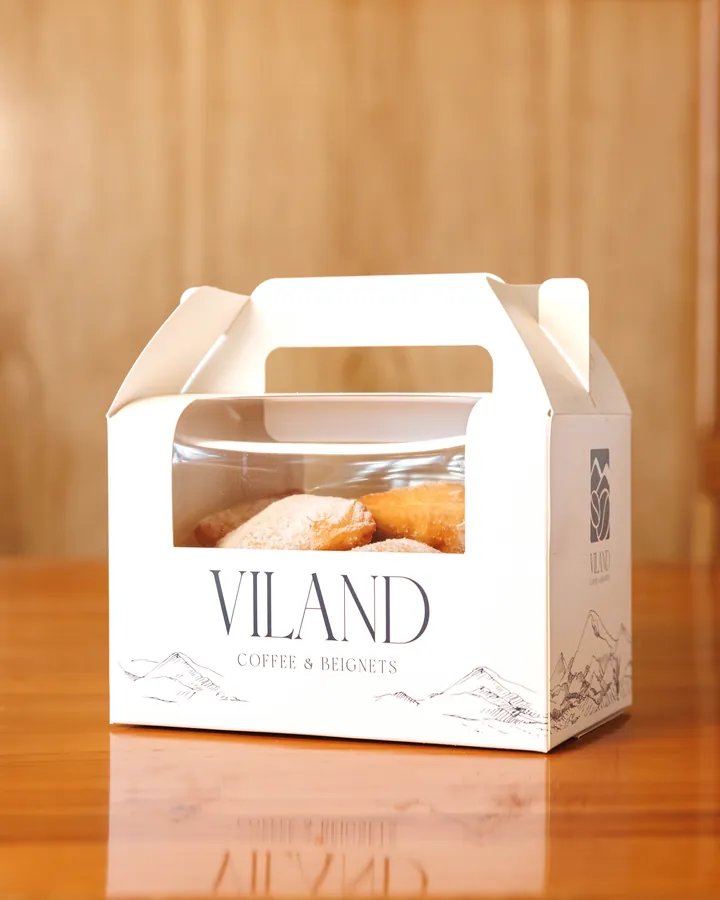 Hộp giấy ViLand có quai xách và ô cửa trong, bên trong là beignet, đặt trên bàn gỗ.