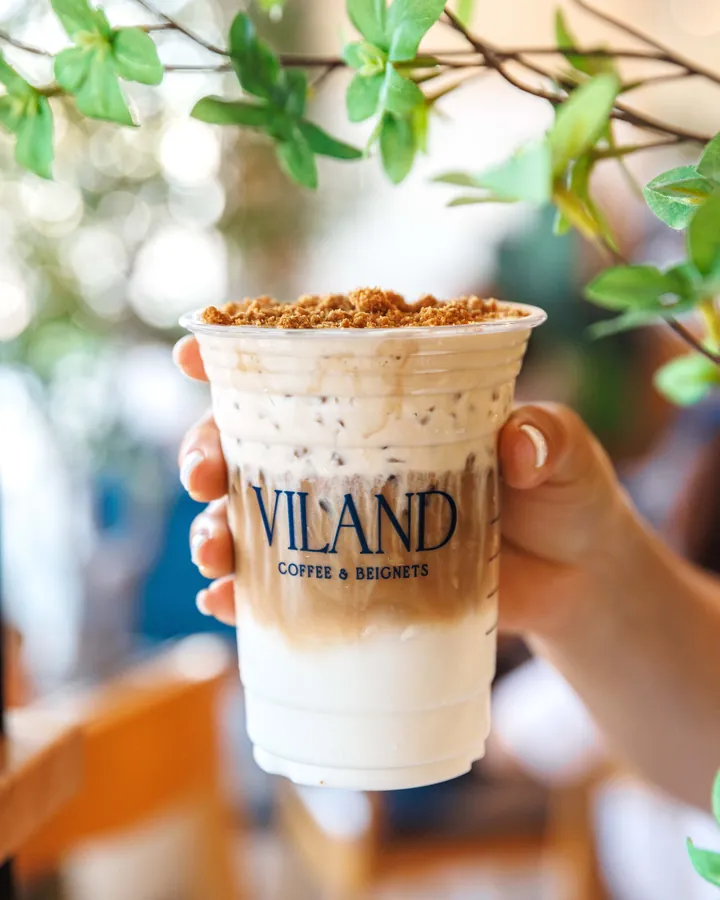 Một bàn tay nâng ly cà phê đá ViLand phủ kem bên trong quán, phía sau là ghế gỗ và lá xanh.