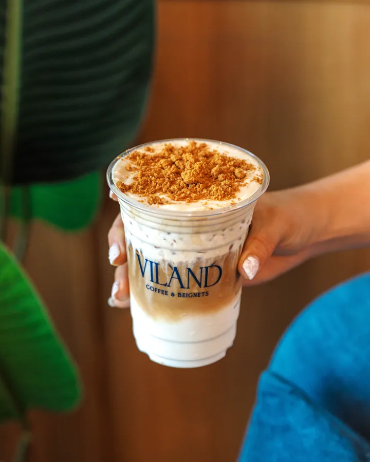 A hand holds out a ViLand iced coffee to go, topped with cream and crumble.