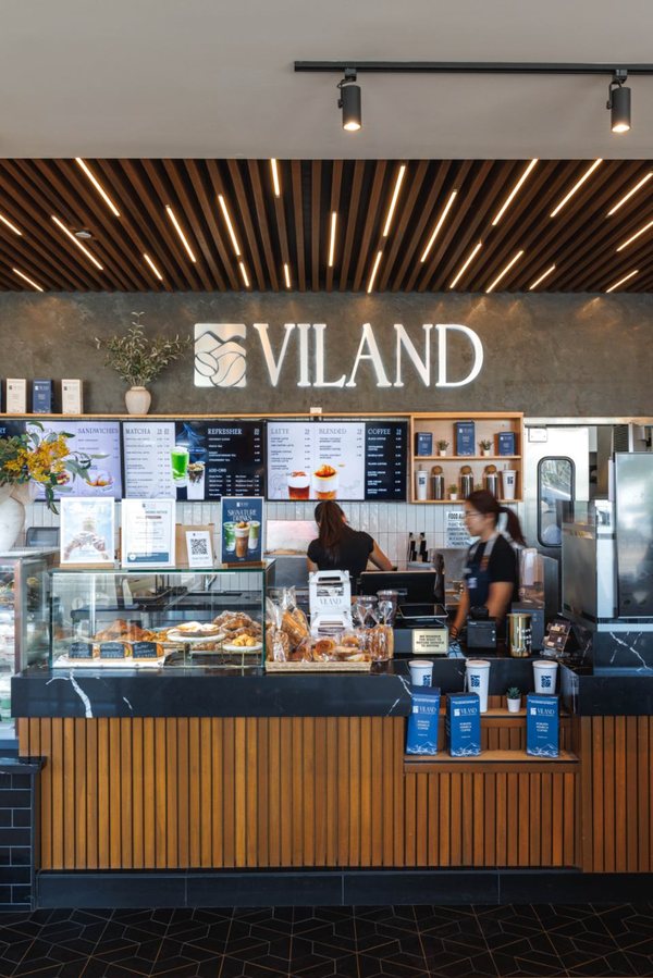 Quầy ViLand với bảng hiệu VILAND phát sáng trên tường đá, trần gỗ ốp thanh, bảng menu, tủ bánh và hai nhân viên đang làm việc sau quầy ốp gỗ.