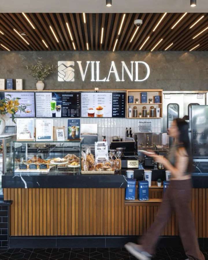 Quầy ViLand dưới trần ốp gỗ, có bảng hiệu ViLand phát sáng, màn hình menu và tủ bánh, một cô gái mặc quần nâu đang đi ngang qua.