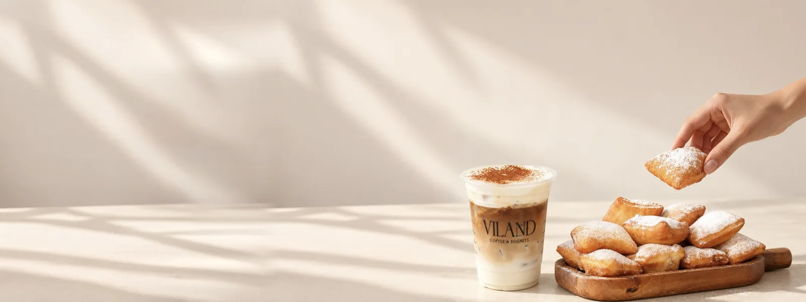 Ly cà phê đá ViLand phủ bột quế bên khay gỗ đầy beignet phủ đường bột, một bàn tay đang nhấc một chiếc lên.