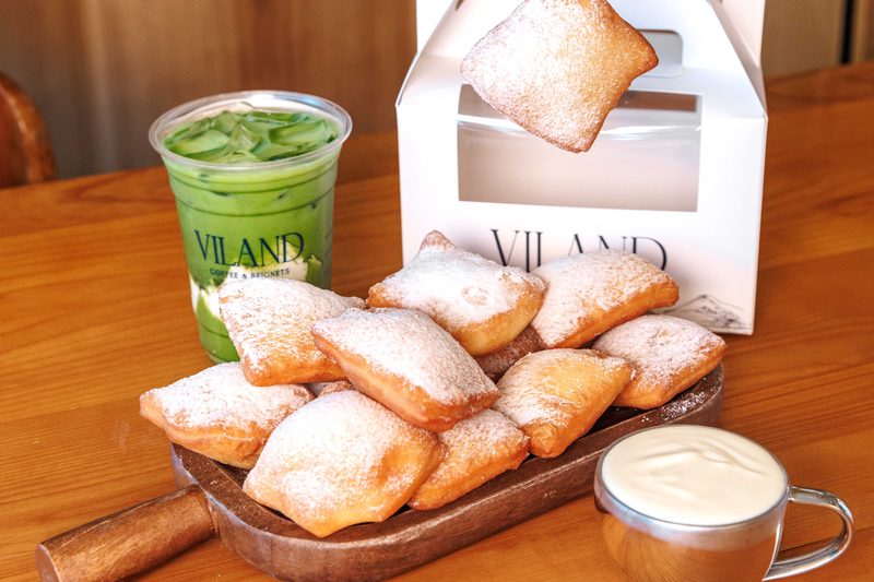 Ly matcha latte đá ViLand bên cạnh tấm thớt gỗ đầy bánh beignet phủ đường bột. Một chiếc bánh được nhấc lên trước hộp mang đi ViLand màu trắng, bên cạnh có ly kem nhỏ.